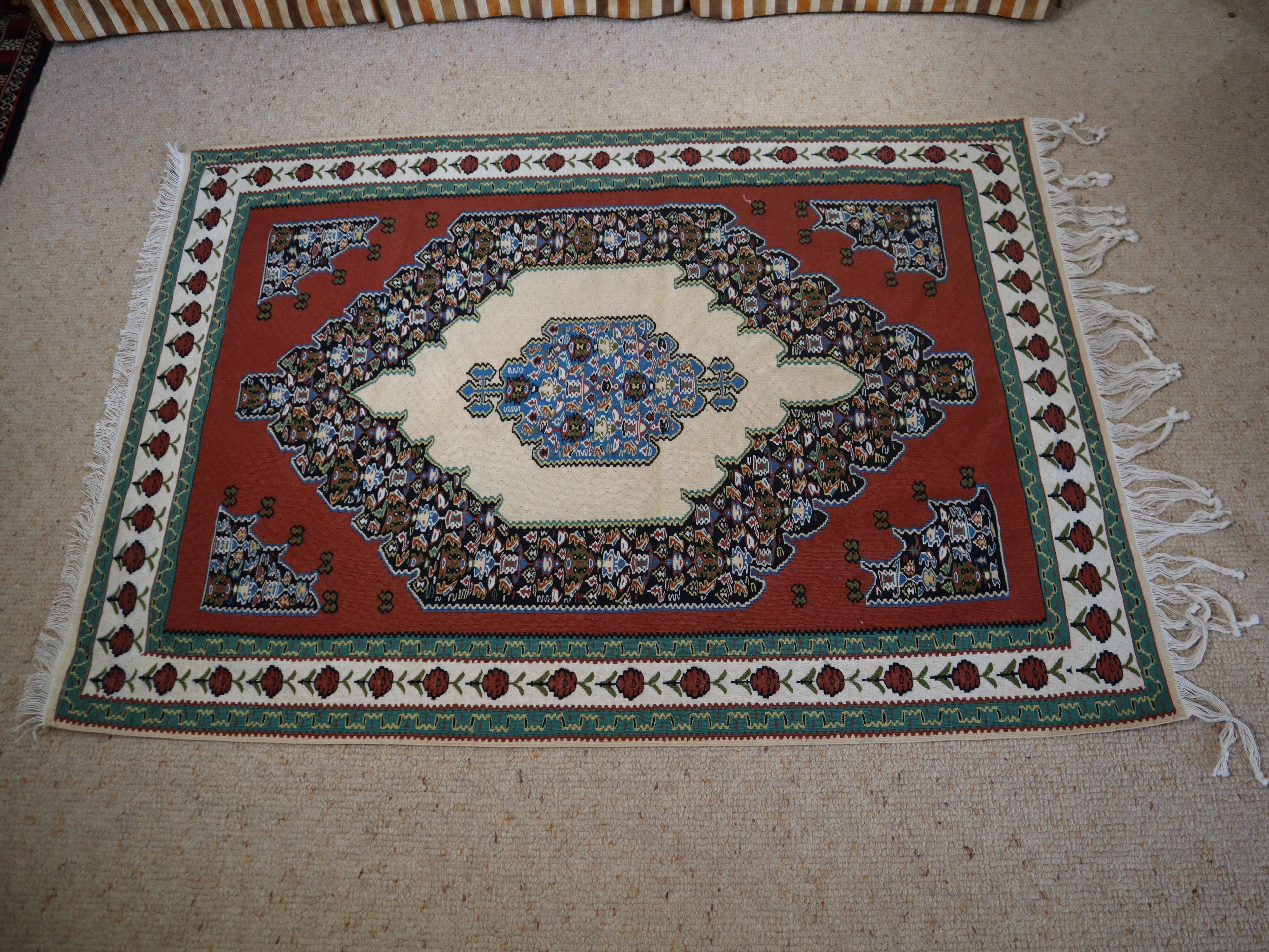 A rectangular woven area rug with a traditional Kurdish-style design, featuring a red field, cream center medallion, and green border, laid flat on a beige carpeted floor.