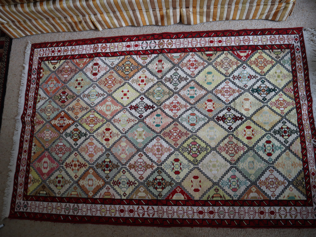 A rectangular patterned rug laid flat on a light-colored floor, featuring a geometric diamond design in cream, red, blue, green, and brown tones, with a striped textile visible at the top edge.