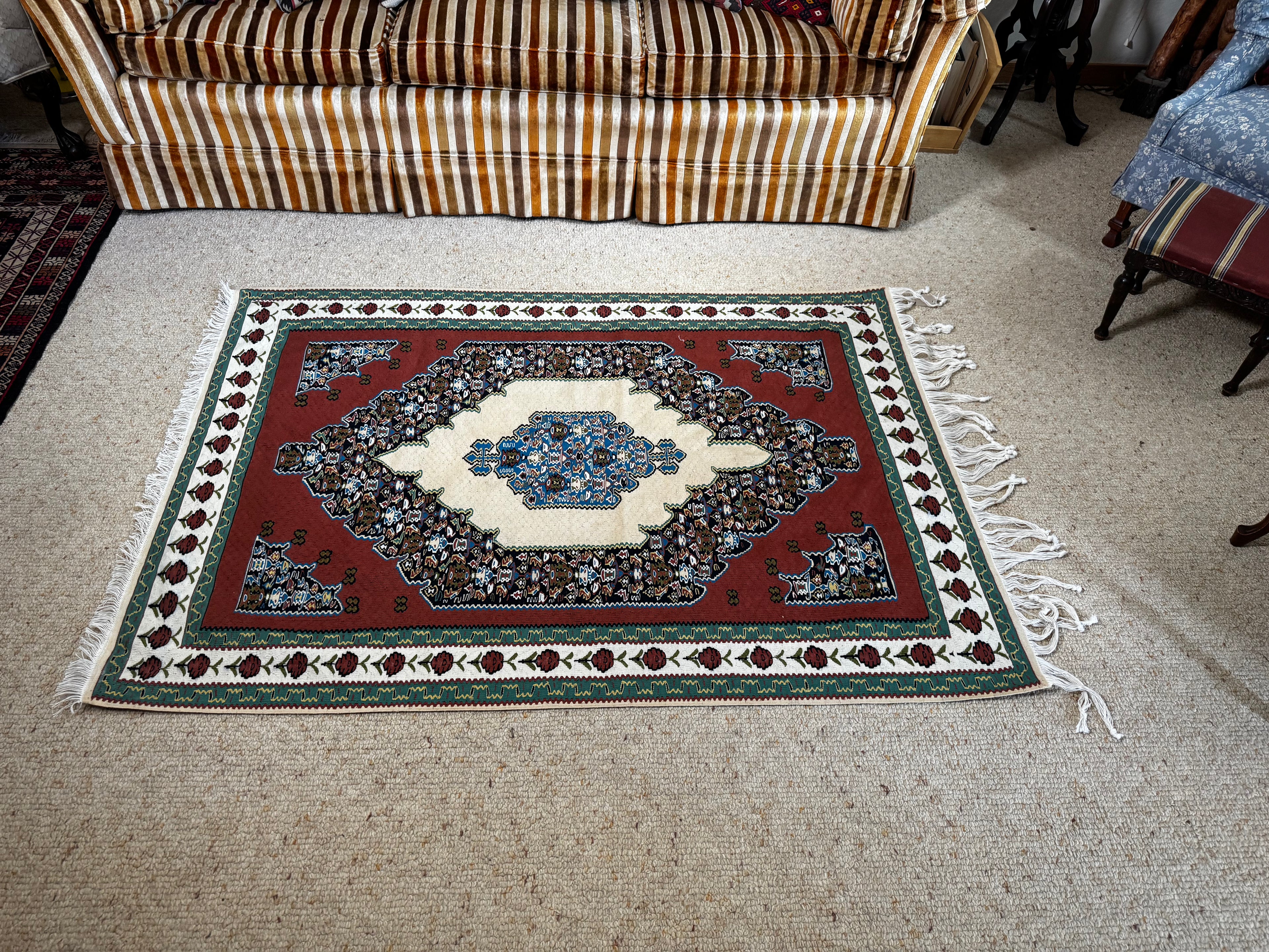 A rectangular Sine area rug with a red central field, cream medallion, and multicolor floral border, laid out on a beige carpeted floor in a living room with a striped sofa visible in the background.