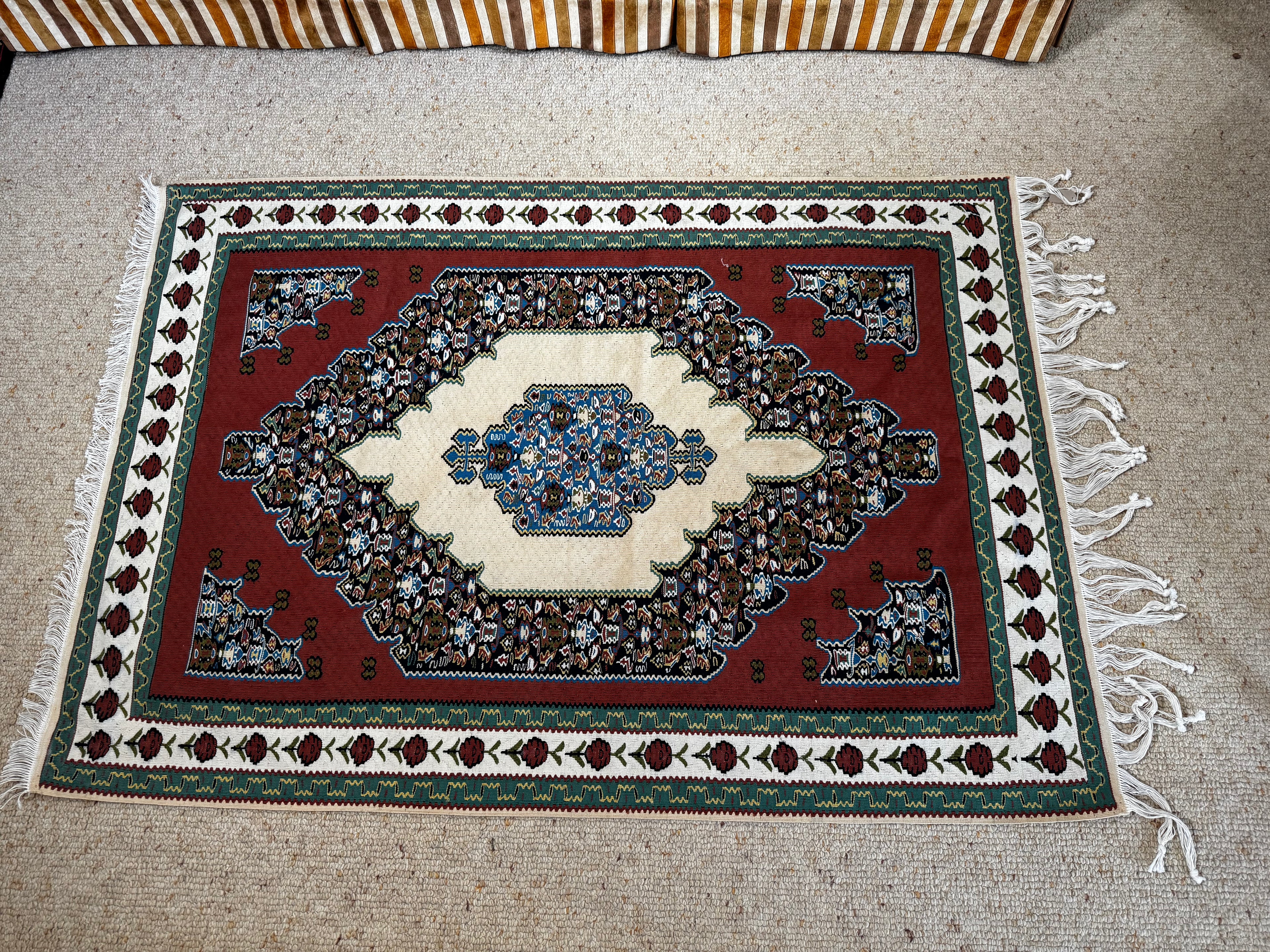 A rectangular traditional  Sine rug with a red field, cream center medallion, and detailed blue, green, and brown border motifs, laid flat on a beige carpeted floor.