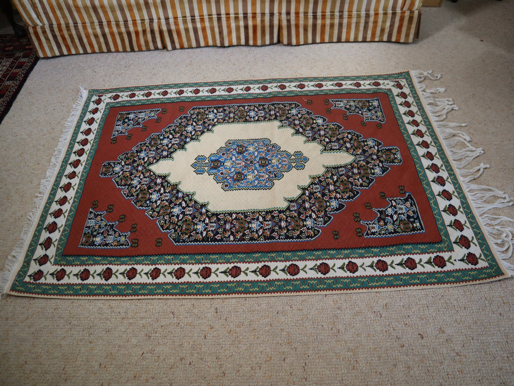 A rectangular Persian-style area rug with a red field, cream center, and ornate floral motifs, laid out on a beige carpet with part of a striped sofa visible at the top.